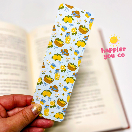 Decorative bookmark with lemon and basket pattern held over an open book, with 'happier you co' branding.
