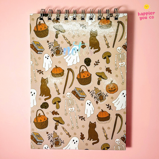A spiral-bound notebook with a beige cover decorated with a Halloween-themed pattern of ghosts, skeletons, pumpkins, mushrooms, and cats, set against a solid pink background.