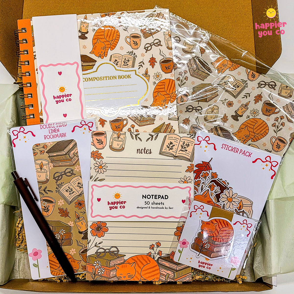 A stationery gift set from happier you co. arranged in a box, featuring a composition book, notepad, sticky notes, sticker pack, and pens with an autumn-themed illustration of books, coffee cups, and an orange cat.