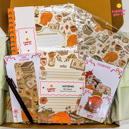 A stationery gift set from happier you co. arranged in a box, featuring a composition book, notepad, sticky notes, sticker pack, and pens with an autumn-themed illustration of books, coffee cups, and an orange cat.
