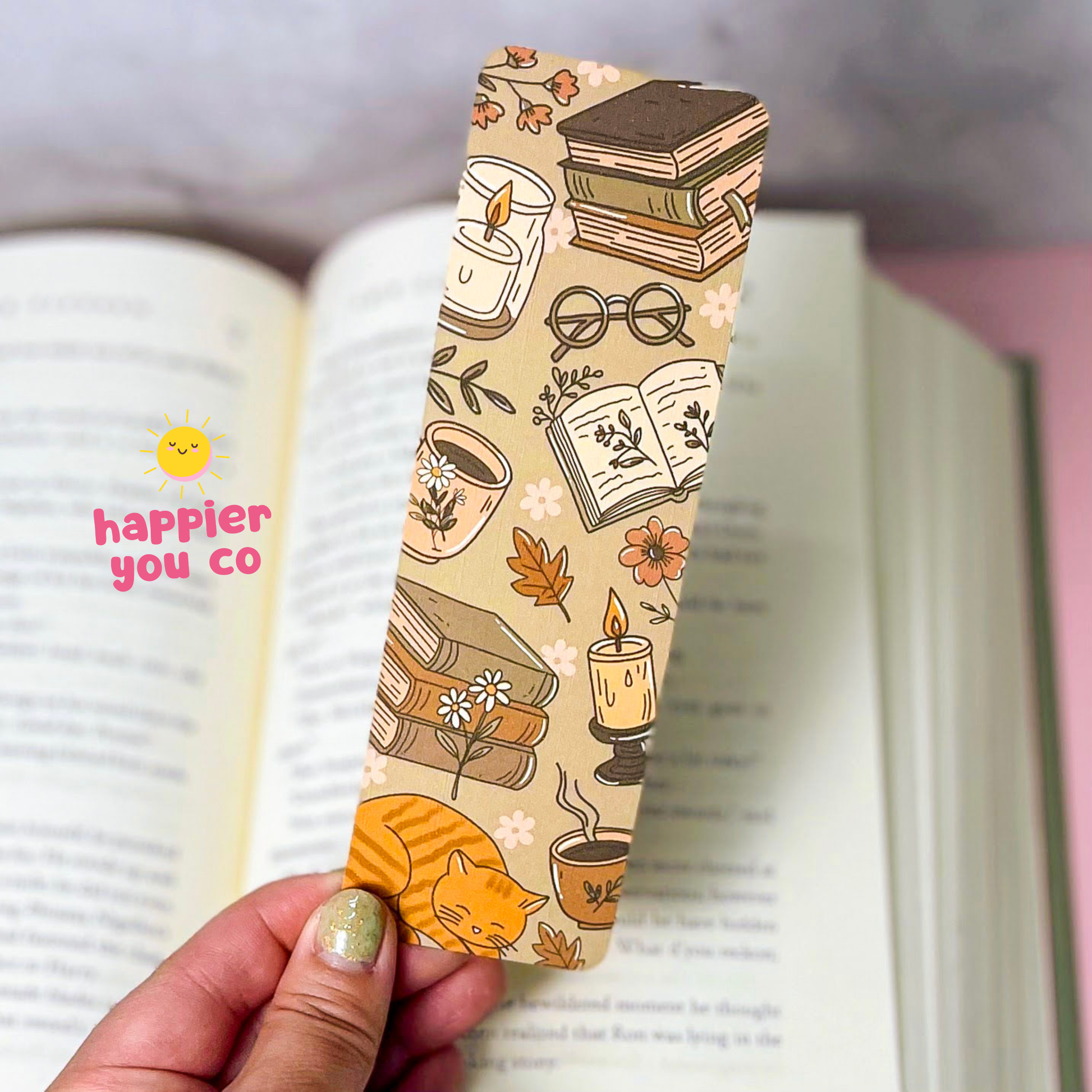 A hand holds a rectangular paper bookmark printed with cozy illustrations of books, candles, glasses, flowers, leaves, and an orange cat. The bookmark is shown over an open book, with a pink background and a small 'happier you co' logo on the left.