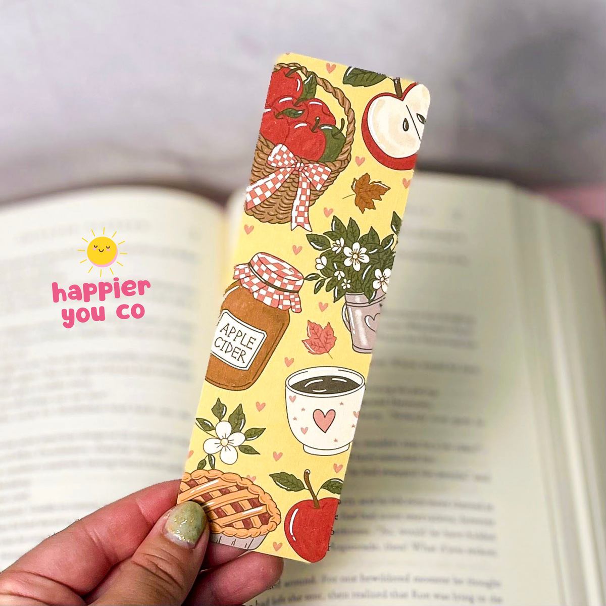 A hand holds a yellow paper bookmark printed with autumn and apple-themed illustrations, including apples, a jar of apple cider, a cup, a pie, and flowers, in front of an open book. The bookmark has a pink gingham ribbon graphic and a small heart at the bottom.