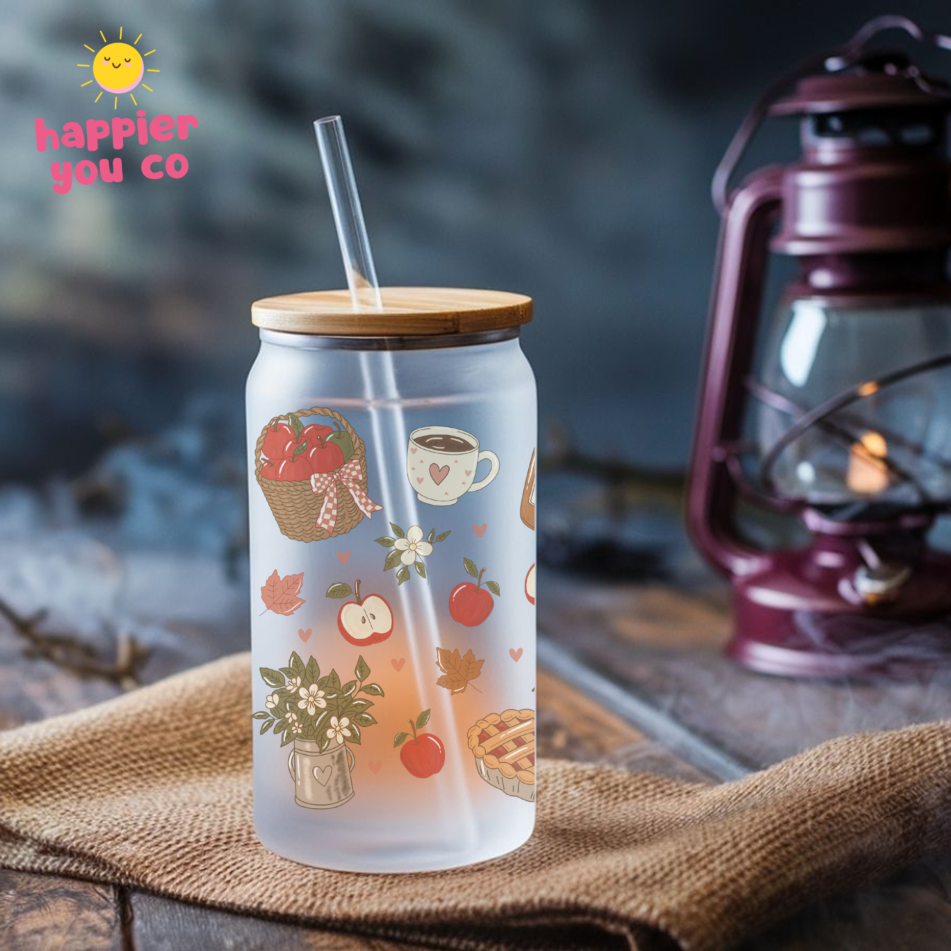 A frosted glass can cooler with a bamboo lid and clear straw, decorated with autumn-themed illustrations like apples, a basket, and a teacup. It is set on a burlap cloth with a rustic lantern in the background.