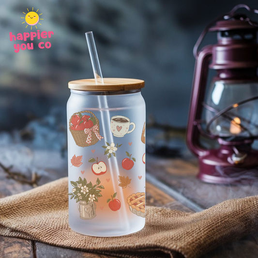 A frosted glass can cooler with a bamboo lid and clear straw, decorated with autumn-themed illustrations like apples, a basket, and a teacup. It is set on a burlap cloth with a rustic lantern in the background.