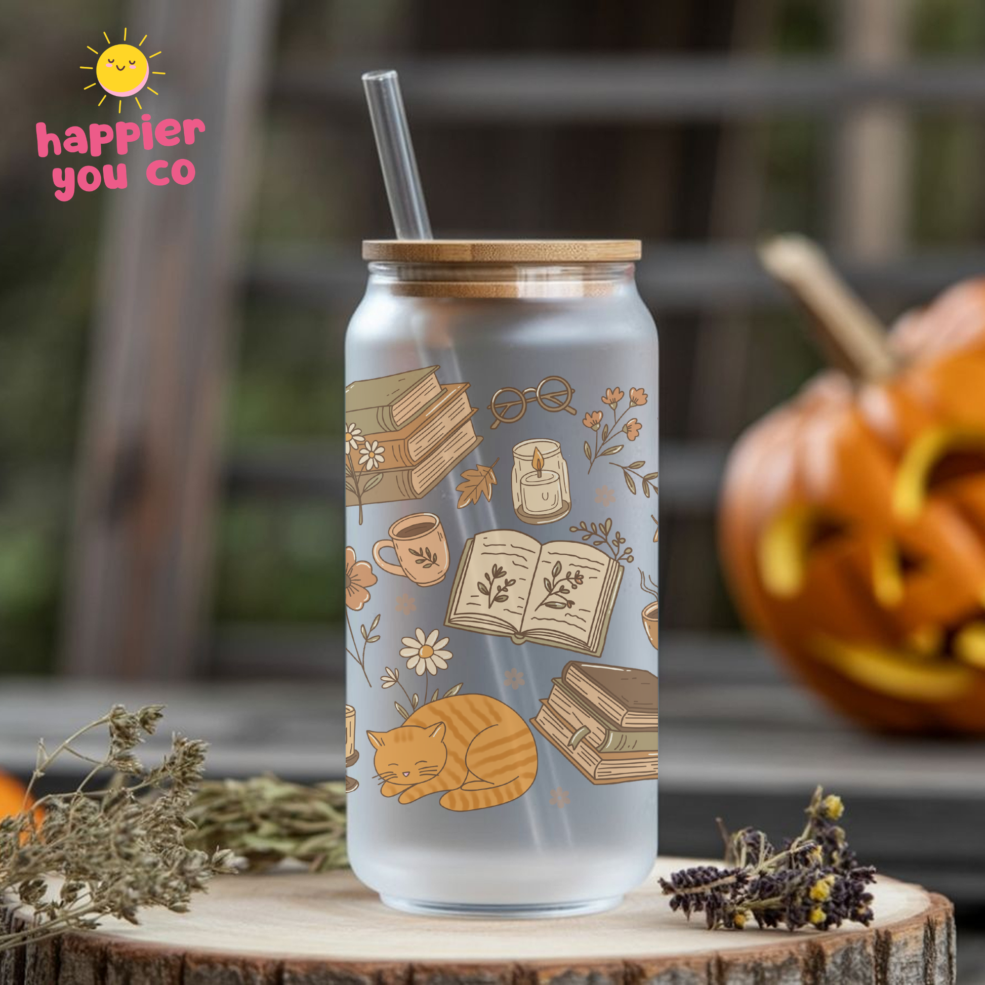 A frosted glass can-style tumbler with a bamboo lid and clear straw, decorated with autumn book-and-cat illustrations. It sits on a wooden surface outdoors with a blurred pumpkin and dried flowers in the background.