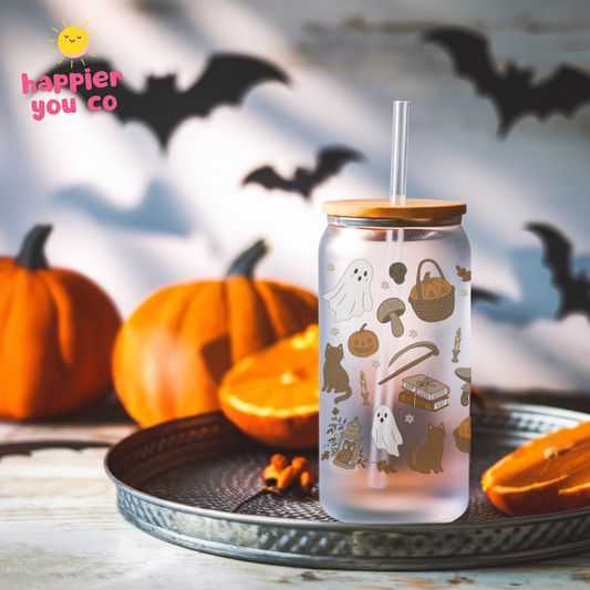 A frosted glass tumbler with a bamboo lid and clear straw, decorated with cute Halloween illustrations including ghosts, cats, pumpkins, and a witch hat. The tumbler is displayed on a metal tray with pumpkins and orange slices in the background, with a 'happier you co' logo in the top-left corner.