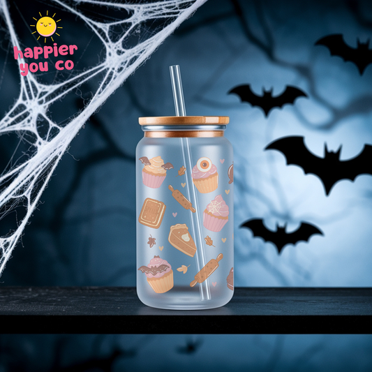 A clear glass can-style tumbler with a bamboo lid and clear straw, decorated with cute cupcake and pastry illustrations. It is set on a dark surface against a spooky Halloween background with cobwebs, bats, and a blue sky.