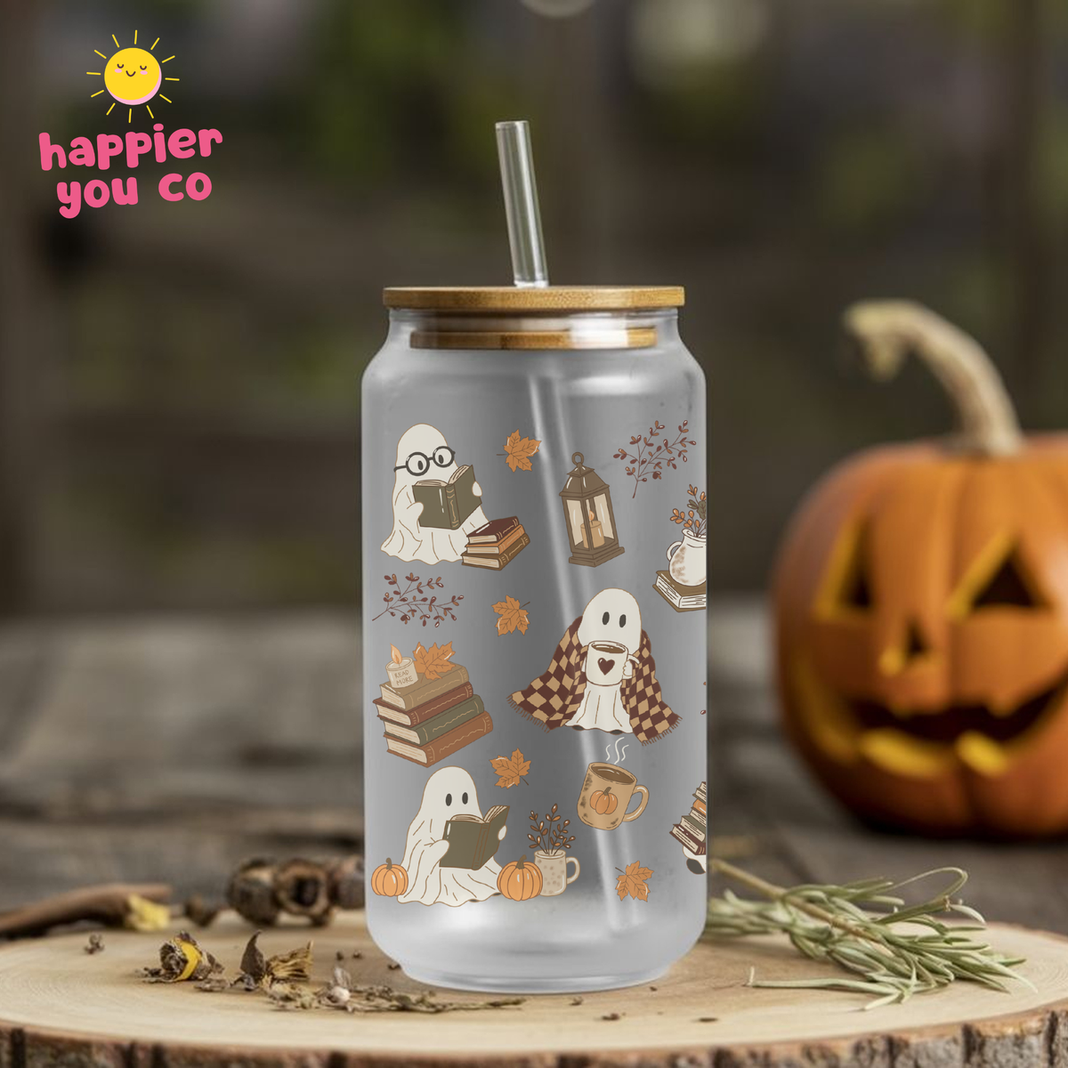 A frosted glass can-shaped tumbler with a bamboo lid and clear straw, decorated with a cute autumn ghost pattern. The tumbler is displayed on a rustic wood slice with a carved jack-o'-lantern and dried flowers in the background, creating a cozy fall atmosphere.