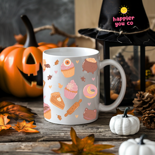 A white ceramic mug with a cute autumn-themed pattern of cupcakes, donuts, a slice of cake, a cauldron, and leaves, styled on a rustic wooden table with pumpkins, autumn leaves, and a witch hat lamp.