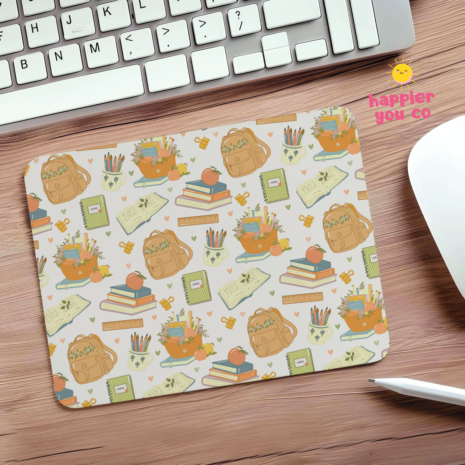 A rectangular desk mat with a cute school-themed pattern of backpacks, books, pencils, and notebooks on a cream background, placed on a wooden desk next to a keyboard, mouse, and stylus.
